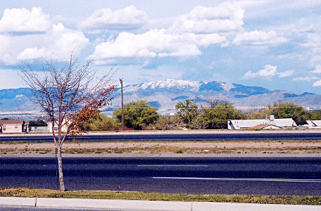 [The pavement of a multilane road is in the foreground with houses and trees in the middle ground. In the background are snow-capped mountains with green tree-covered sections. There are lots of puffy clouds in the sky.]